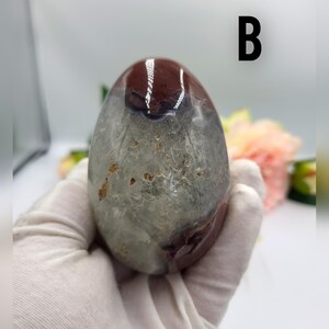 May include: A polished, oval-shaped gemstone with a mix of colors. The top portion is a deep burgundy, transitioning to a translucent gray-green lower section. The letter "B" is printed in black on the background.