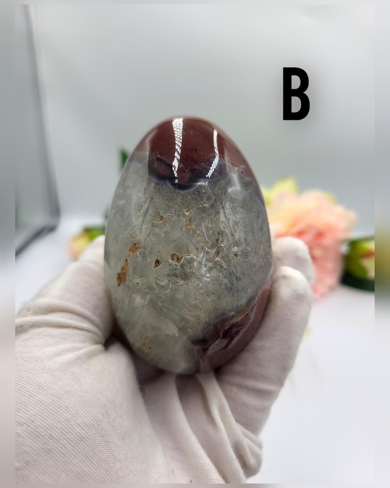 May include: A polished, oval-shaped gemstone with a mix of colors. The top portion is a deep burgundy, transitioning to a translucent gray-green lower section. The letter "B" is printed in black on the background.