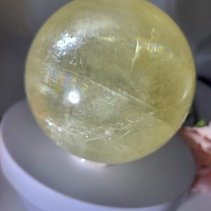 May include: A large, smooth, yellow crystal ball. The ball is made of clear quartz with a slight yellow tint. The ball is sitting on a white stand.
