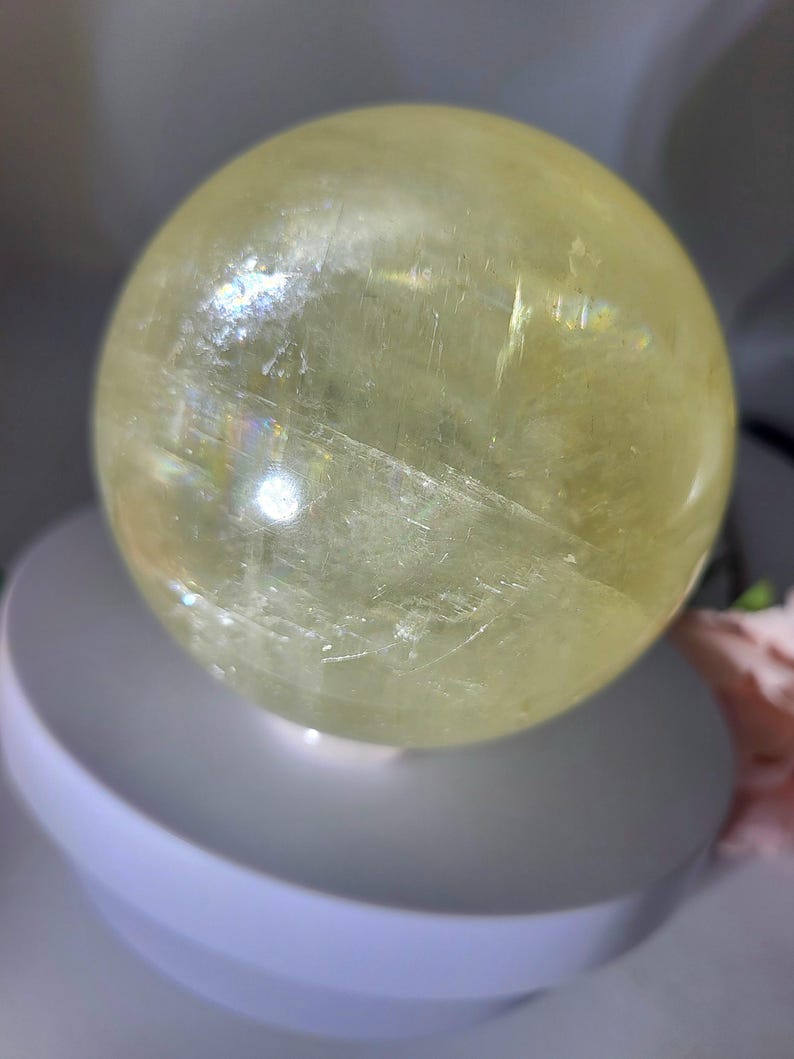 May include: A large, smooth, yellow crystal ball. The ball is made of clear quartz with a slight yellow tint. The ball is sitting on a white stand.