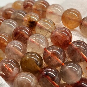 May include: Close-up of a collection of clear, round beads with internal reddish-brown inclusions. The beads are strung together with orange string, creating a natural, earthy aesthetic. The beads are likely used for jewelry making.