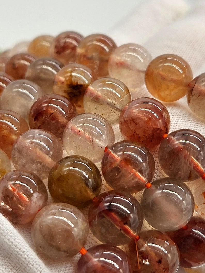 May include: Close-up of a collection of clear, round beads with internal reddish-brown inclusions. The beads are strung together with orange string, creating a natural, earthy aesthetic. The beads are likely used for jewelry making.