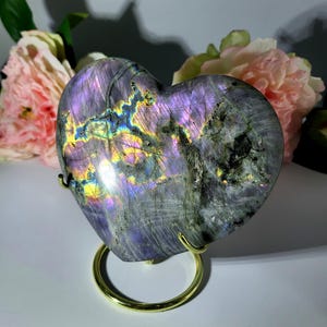 May include: A heart-shaped labradorite stone with iridescent purple, blue, and yellow flashes. The polished stone is displayed on a gold-colored metal stand, with pink flowers in the background. The stone's surface has a textured appearance.