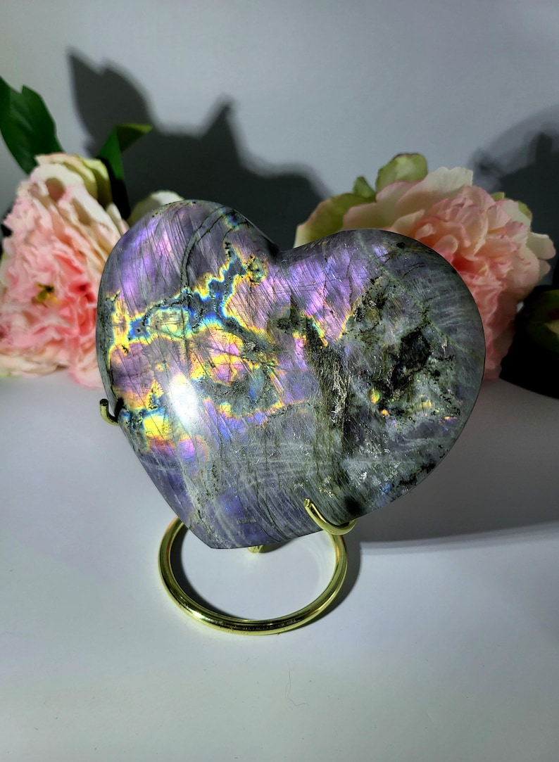 May include: A heart-shaped labradorite stone with iridescent purple, blue, and yellow flashes. The polished stone is displayed on a gold-colored metal stand, with pink flowers in the background. The stone's surface has a textured appearance.