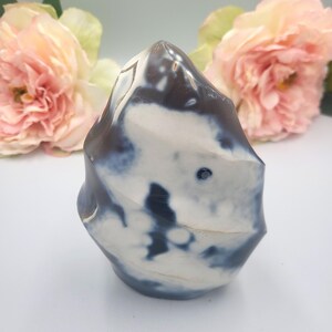 May include: A decorative, polished stone sculpture with a unique, organic shape. The stone features a blend of white, dark blue, and brown colors. The sculpture is displayed against a soft, blurred background with pink flowers.