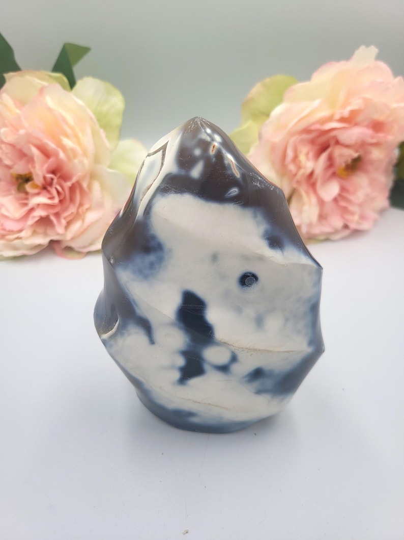 May include: A decorative, polished stone sculpture with a unique, organic shape. The stone features a blend of white, dark blue, and brown colors. The sculpture is displayed against a soft, blurred background with pink flowers.