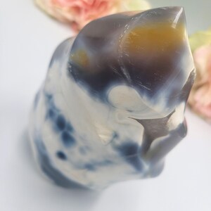 May include: A polished, abstract-shaped stone with a mix of colors. The stone features swirling patterns of white, navy blue, and a touch of yellow-orange. The top of the stone has a smooth, rounded shape. The background includes soft pink and yellow flowers.