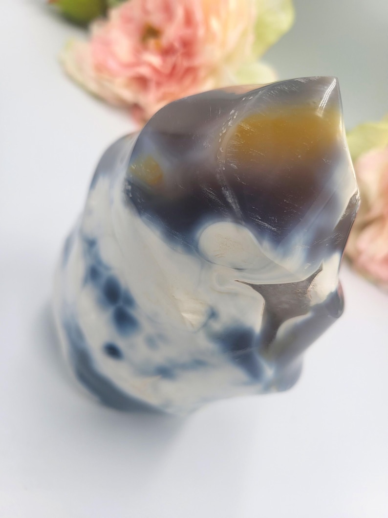 May include: A polished, abstract-shaped stone with a mix of colors. The stone features swirling patterns of white, navy blue, and a touch of yellow-orange. The top of the stone has a smooth, rounded shape. The background includes soft pink and yellow flowers.