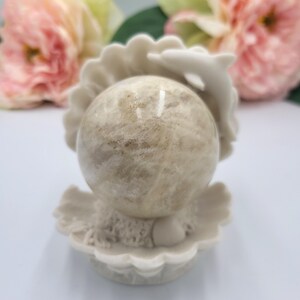 May include: A beige sphere rests in a white seashell-shaped holder with a dolphin accent. The sphere has a mottled appearance, and the holder has a textured surface. Pink and yellow flowers are in the background, adding a soft, natural touch.
