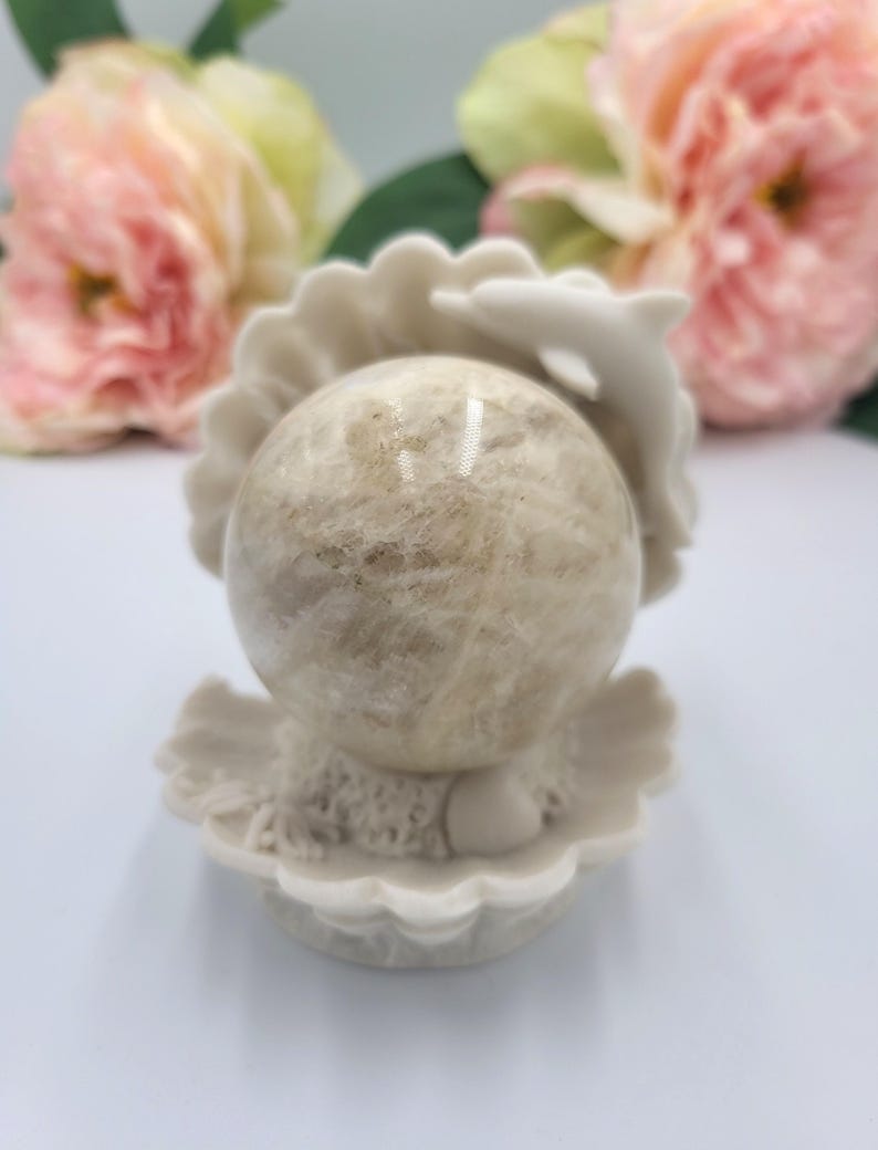 May include: A beige sphere rests in a white seashell-shaped holder with a dolphin accent. The sphere has a mottled appearance, and the holder has a textured surface. Pink and yellow flowers are in the background, adding a soft, natural touch.