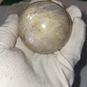 May include: A polished, spherical moonstone. The stone has a creamy white base color with iridescent flashes of blue and gold. The sphere is held in a white gloved hand, with a soft focus background of pink flowers.