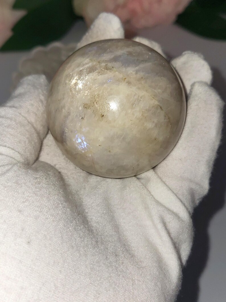 May include: A polished, spherical moonstone. The stone has a creamy white base color with iridescent flashes of blue and gold. The sphere is held in a white gloved hand, with a soft focus background of pink flowers.