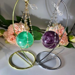 May include: Two crystal spheres, one green and one purple, are suspended from gold and silver metal stands. Each sphere is adorned with a delicate chain of beads and a small decorative charm.