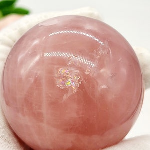 May include: A large, pink rose quartz sphere with iridescent inclusions. The sphere is smooth and polished, and it is resting on a white surface.
