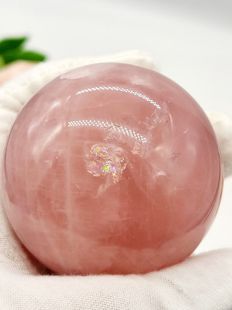 May include: A large, pink rose quartz sphere with iridescent inclusions. The sphere is smooth and polished, and it is resting on a white surface.