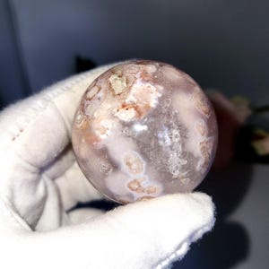 May include: A spherical gemstone with a translucent, milky appearance. The sphere has a pattern of light brown and white inclusions, creating a unique, natural design. The gemstone is held in a white glove.