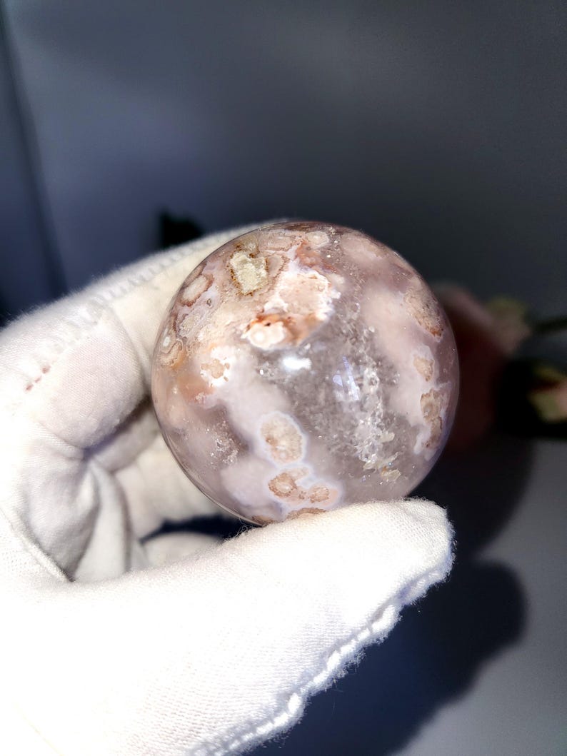 May include: A spherical gemstone with a translucent, milky appearance. The sphere has a pattern of light brown and white inclusions, creating a unique, natural design. The gemstone is held in a white glove.