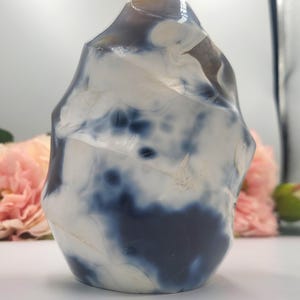 May include: A polished, freeform stone sculpture with a marbled appearance. The stone features a mix of white and deep blue hues, creating a striking contrast. The sculpture's shape is organic and irregular, with a smooth, glossy finish. The background includes pink flowers.