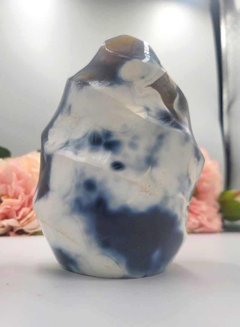 May include: A polished, freeform stone sculpture with a marbled appearance. The stone features a mix of white and deep blue hues, creating a striking contrast. The sculpture's shape is organic and irregular, with a smooth, glossy finish. The background includes pink flowers.