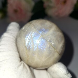 May include: A round, off-white moonstone sphere with iridescent blue and white flashes. The sphere is held in a white gloved hand, with soft pink flowers in the background. The moonstone is a natural gemstone.