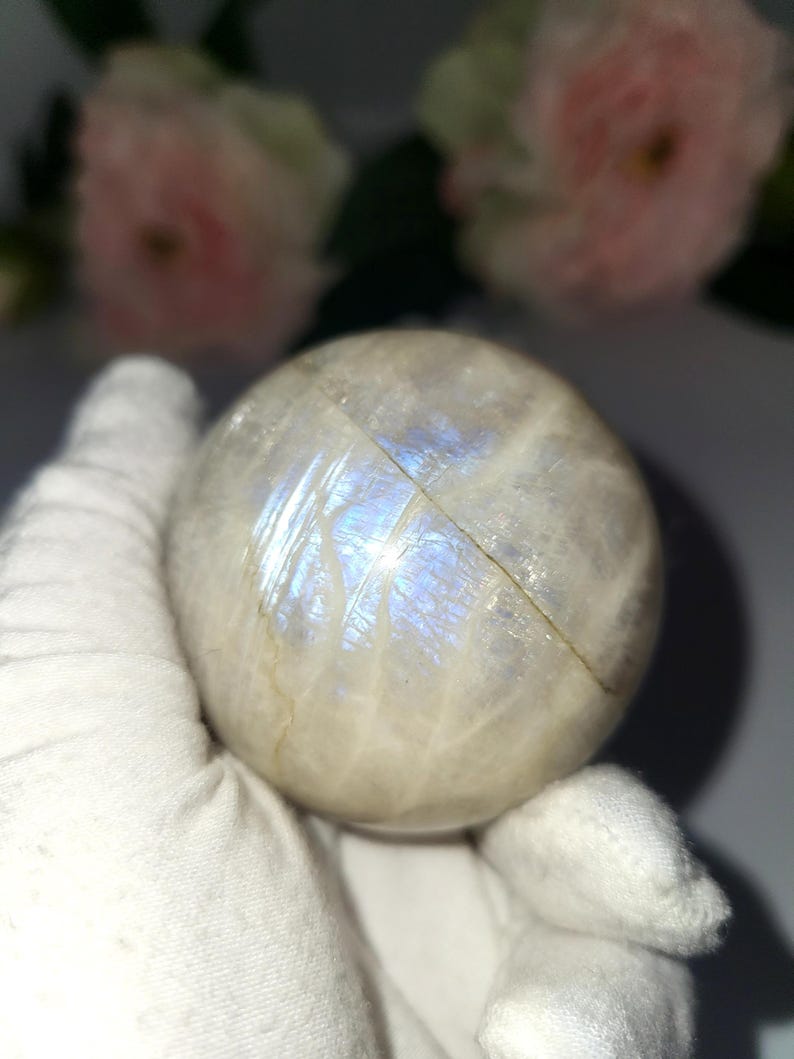 May include: A round, off-white moonstone sphere with iridescent blue and white flashes. The sphere is held in a white gloved hand, with soft pink flowers in the background. The moonstone is a natural gemstone.