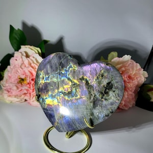 May include: A heart-shaped labradorite stone with iridescent colors of purple, blue, and yellow, displayed on a gold-colored metal stand. The stone is set against a backdrop of pink flowers, creating a visually appealing contrast.