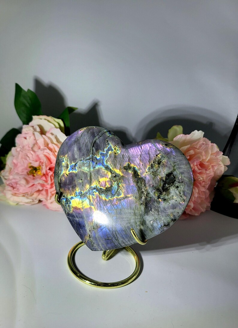 May include: A heart-shaped labradorite stone with iridescent colors of purple, blue, and yellow, displayed on a gold-colored metal stand. The stone is set against a backdrop of pink flowers, creating a visually appealing contrast.