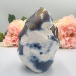 May include: A polished stone sculpture with a rounded, irregular shape. The stone features a mix of white, dark blue, and brown colors. The sculpture is set against a soft, blurred background with pink flowers.