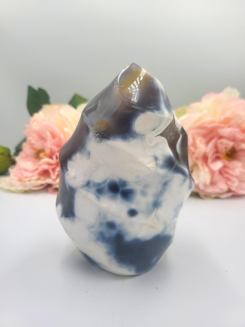 May include: A polished stone sculpture with a rounded, irregular shape. The stone features a mix of white, dark blue, and brown colors. The sculpture is set against a soft, blurred background with pink flowers.