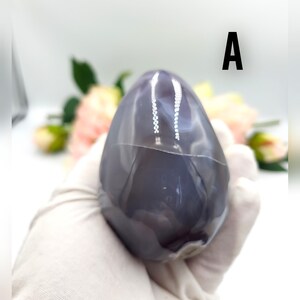 May include: A polished, egg-shaped stone with a smooth, glossy surface. The stone is primarily a deep gray color with lighter gray and white veining. The letter "A" is printed in black on the upper right side of the image. The stone is held by a gloved hand.