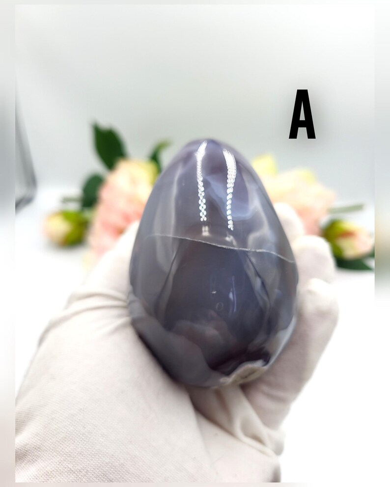 May include: A polished, egg-shaped stone with a smooth, glossy surface. The stone is primarily a deep gray color with lighter gray and white veining. The letter "A" is printed in black on the upper right side of the image. The stone is held by a gloved hand.