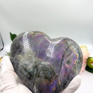 May include: A heart-shaped labradorite stone with a polished surface. The stone displays a mix of gray, purple, and iridescent colors. The heart is held by a gloved hand, with a blurred floral background.