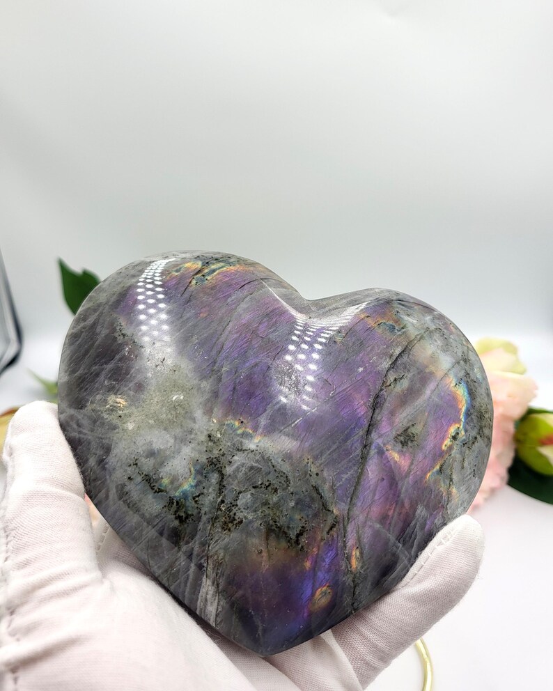 May include: A heart-shaped labradorite stone with a polished surface. The stone displays a mix of gray, purple, and iridescent colors. The heart is held by a gloved hand, with a blurred floral background.