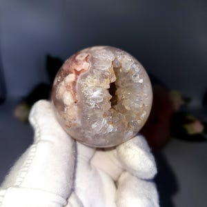 May include: A spherical geode with a rough, crystalline interior and a smooth, translucent exterior. The geode's surface displays a mix of pink and white hues, with a visible cavity revealing the crystal structure within. The object is held by a gloved hand.