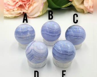 Op de afbeelding: Vijf blauwe kantagaat bollen, elk gemonteerd op een heldere standaard, worden getoond tegen een achtergrond van roze en crèmekleurige bloemen. De bollen hebben een gestreepte uitstraling met verschillende tinten blauw en wit.