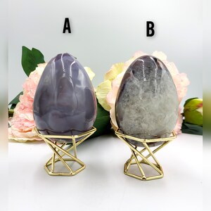 May include: Two polished stone eggs, one purple and one gray, displayed on gold geometric stands. The eggs have a smooth, glossy finish and are set against a backdrop of pink and yellow flowers. The letter "A" is above the purple egg and the letter "B" is above the gray egg.