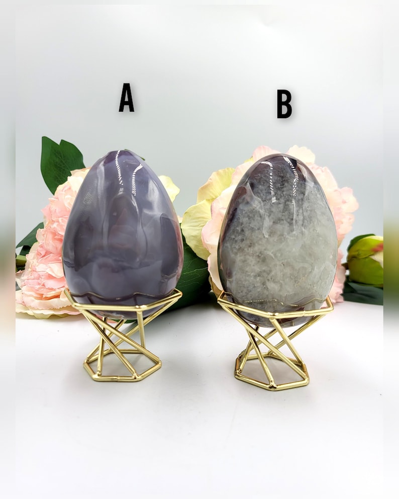 May include: Two polished stone eggs, one purple and one gray, displayed on gold geometric stands. The eggs have a smooth, glossy finish and are set against a backdrop of pink and yellow flowers. The letter "A" is above the purple egg and the letter "B" is above the gray egg.