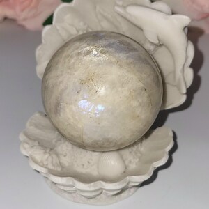May include: A white, seashell-shaped sculpture cradles a pearlescent sphere. A small, white dolphin leaps from the shell. The sphere has iridescent qualities, reflecting light. The sculpture is a decorative item, possibly for a coastal or nautical theme.