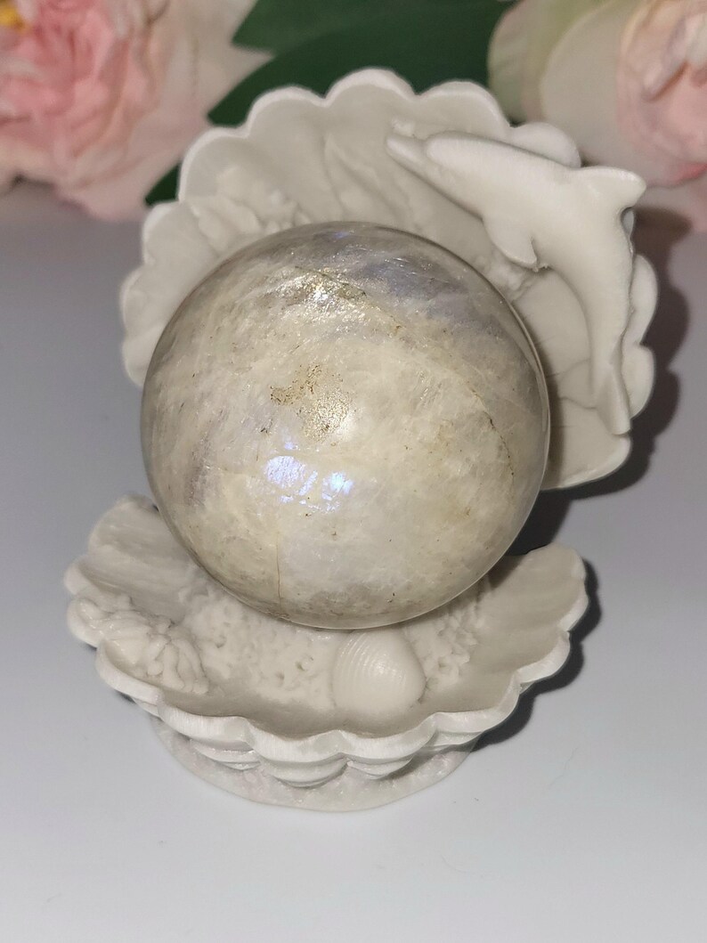 May include: A white, seashell-shaped sculpture cradles a pearlescent sphere. A small, white dolphin leaps from the shell. The sphere has iridescent qualities, reflecting light. The sculpture is a decorative item, possibly for a coastal or nautical theme.