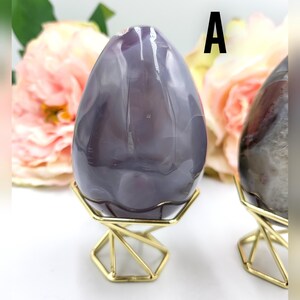 May include: A large, polished, egg-shaped stone in shades of purple and gray, displayed on a gold geometric stand. The stone has natural veining and is set against a backdrop of soft pink flowers. The letter "A" is visible.