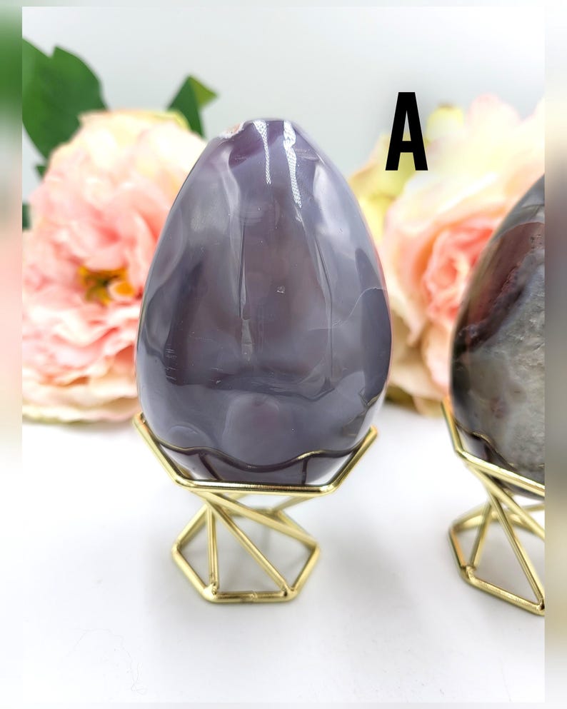 May include: A large, polished, egg-shaped stone in shades of purple and gray, displayed on a gold geometric stand. The stone has natural veining and is set against a backdrop of soft pink flowers. The letter "A" is visible.