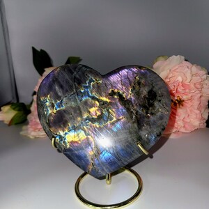 May include: A heart-shaped labradorite stone with iridescent purple, blue, and gold hues. The polished stone is displayed on a gold-colored metal stand. Pink flowers are in the background. A decorative item for home decor or a unique gift.