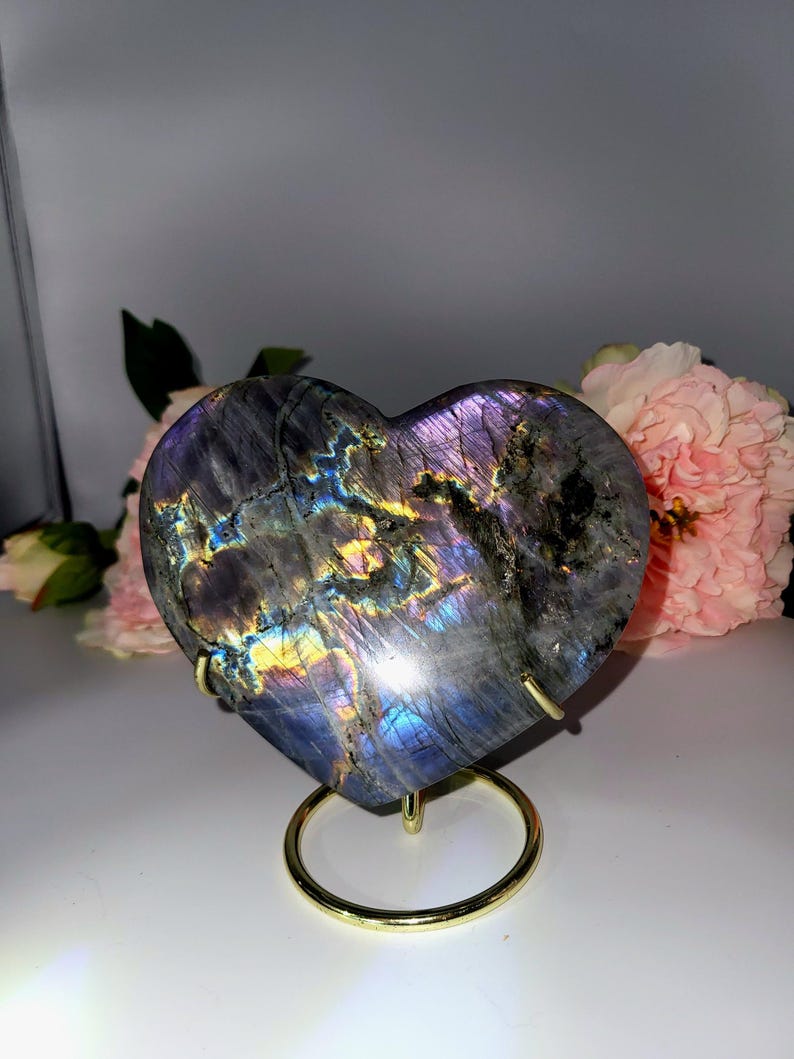 May include: A heart-shaped labradorite stone with iridescent purple, blue, and gold hues. The polished stone is displayed on a gold-colored metal stand. Pink flowers are in the background. A decorative item for home decor or a unique gift.