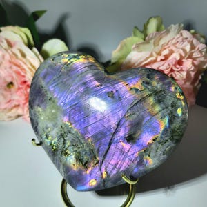 May include: A heart-shaped labradorite stone with iridescent purple, blue, and green hues. The polished stone is displayed on a gold-colored metal stand, with pink flowers in the background. The stone's surface reflects light, creating a shimmering effect.
