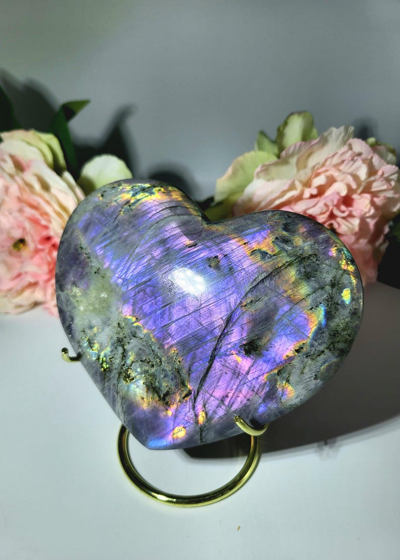 May include: A heart-shaped labradorite stone with iridescent purple, blue, and green hues. The polished stone is displayed on a gold-colored metal stand, with pink flowers in the background. The stone's surface reflects light, creating a shimmering effect.