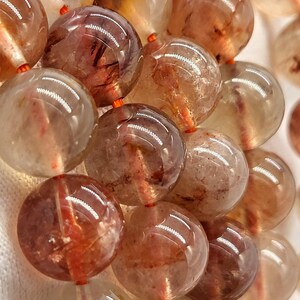 May include: Close-up of a strand of large, round beads. The beads are a translucent, light brown color with reddish-brown inclusions. They are strung on a thin, orange cord. The beads have a polished, glossy finish, reflecting light.