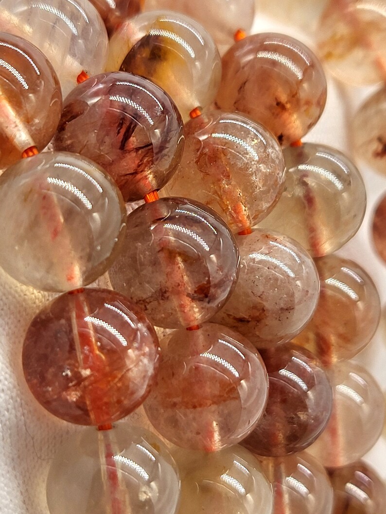 May include: Close-up of a strand of large, round beads. The beads are a translucent, light brown color with reddish-brown inclusions. They are strung on a thin, orange cord. The beads have a polished, glossy finish, reflecting light.