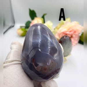 May include: A polished, egg-shaped agate stone in shades of gray and purple, held by a gloved hand. The stone has a smooth, glossy surface with natural patterns and inclusions. The letter "A" is visible in the background.