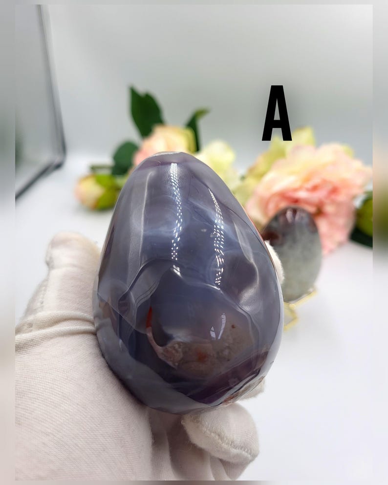 May include: A polished, egg-shaped agate stone in shades of gray and purple, held by a gloved hand. The stone has a smooth, glossy surface with natural patterns and inclusions. The letter "A" is visible in the background.
