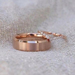 May include: A pair of rose gold wedding bands. The main band is a simple, flat band with a brushed finish. The smaller band is a delicate, curved band with small diamonds set in a V-shape.
